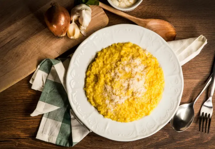 Un cremoso risotto alla milanese, spolverato di parmigiano, pronto per essere gustato. Accanto, cipolla e aglio, ingredienti essenziali.