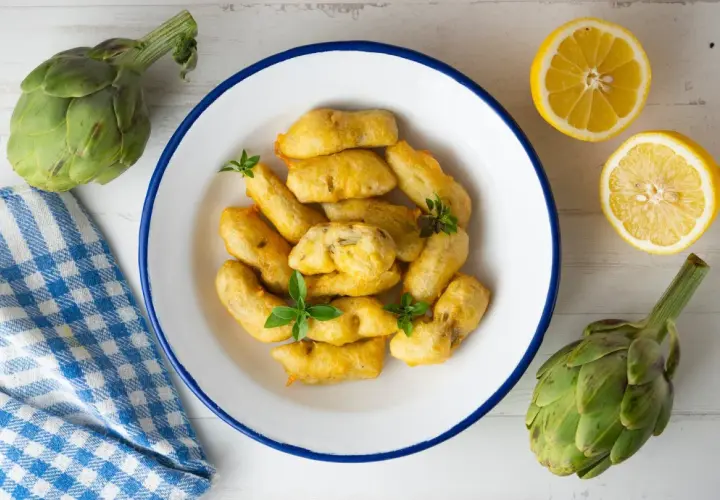 Carciofi fritti dorati, preparati con una pastella croccante, serviti in un piatto bianco con limone e carciofi freschi.