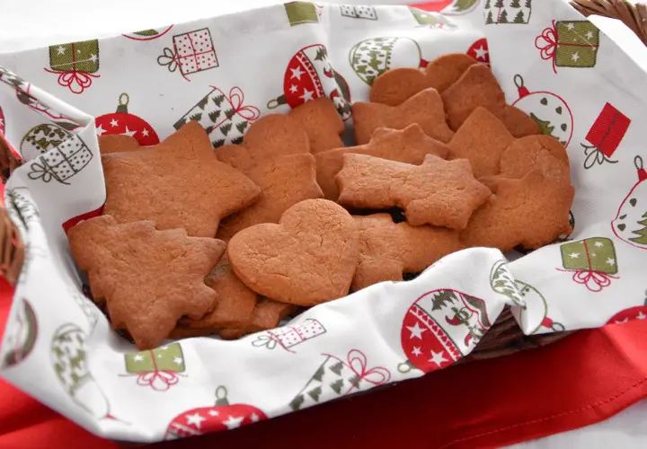 Cesto pieno di biscotti alla cannella a forma di stella, cuore e albero, su un telo natalizio.