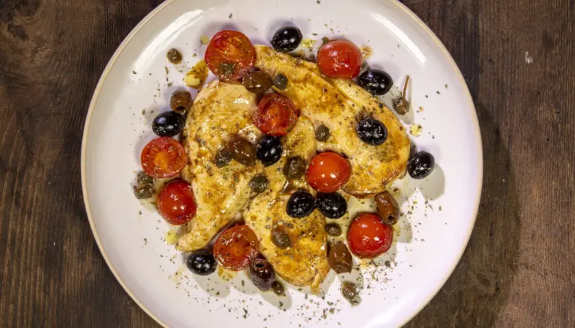 Petto di pollo in padella con pomodorini, olive nere e capperi, condito con erbe aromatiche.