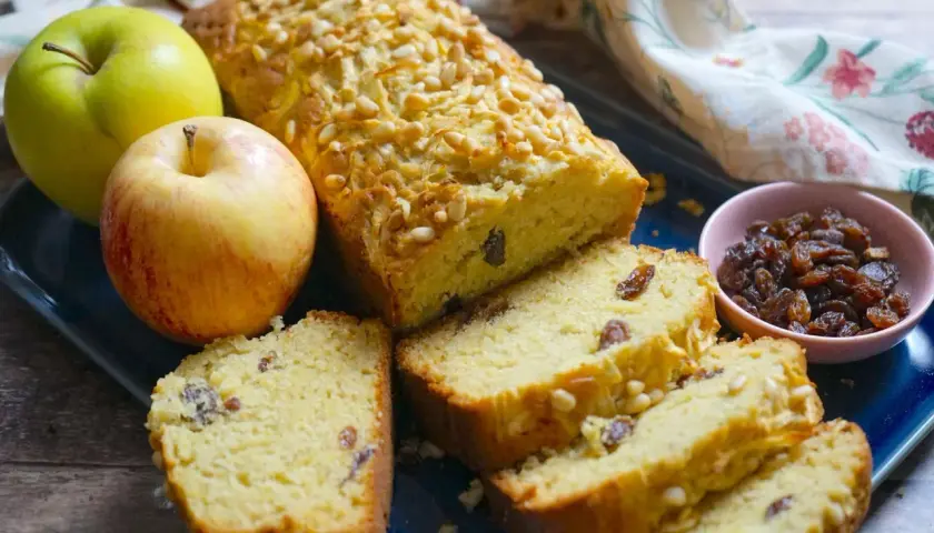 Plumcake alle mele soffice e profumato, con uvetta e pinoli, accompagnato da mele fresche e una ciotolina di uvetta.