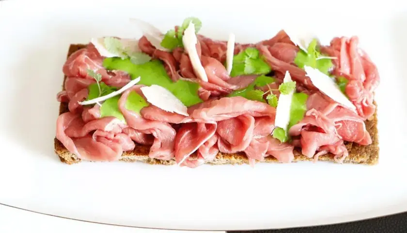 Delicato carpaccio di manzo su crostino integrale, guarnito con crema verde, scaglie di parmigiano e germogli freschi.