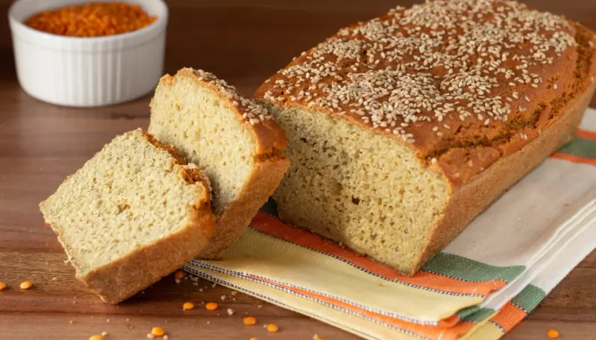 Pane di lenticchie dorato e soffice, cosparso di semi di sesamo, con due fette tagliate. Sullo sfondo, una ciotola di lenticchie rosse.