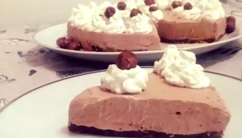Una fetta di golosa cheesecake alla Nutella, guarnita con panna montata e nocciole intere, su un piatto bianco.