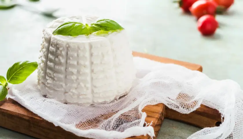 Ricotta fatta in casa, fresca e profumata, guarnita con basilico su un tagliere di legno. Pomodorini rossi sfocati sullo sfondo.