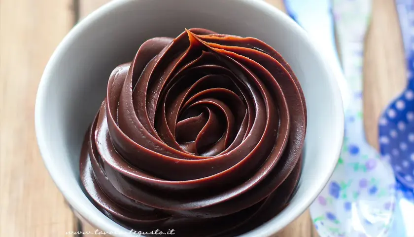 Una ciotola bianca piena di ganache al cioccolato, con un vortice perfetto. Vicino, cucchiai colorati.