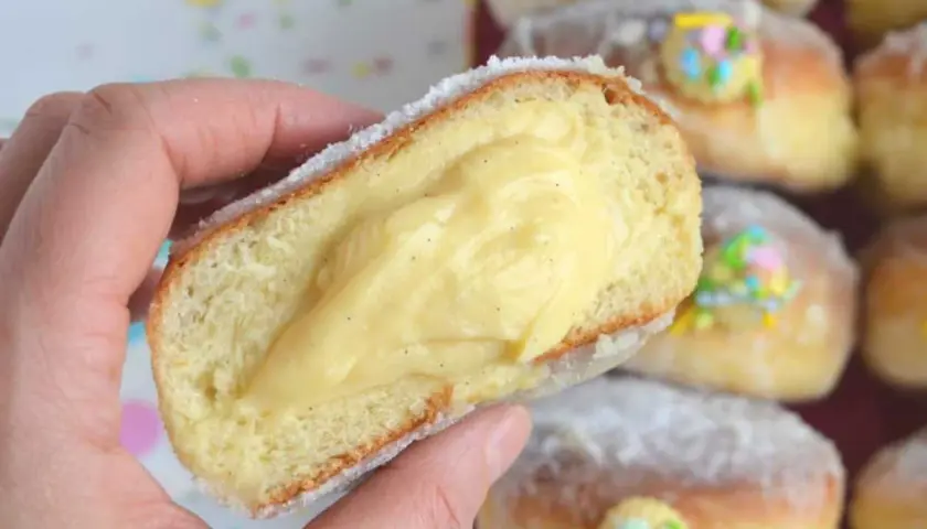 Un bombolone alla crema, soffice e ripieno, viene tenuto in mano. Altri bomboloni alla crema decorati con confettini colorati sono sullo sfondo.