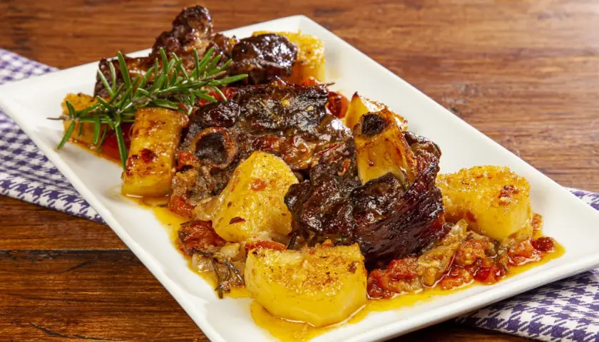 Un succulento agnello al forno con patate dorate, pomodorini e rosmarino su un piatto bianco.