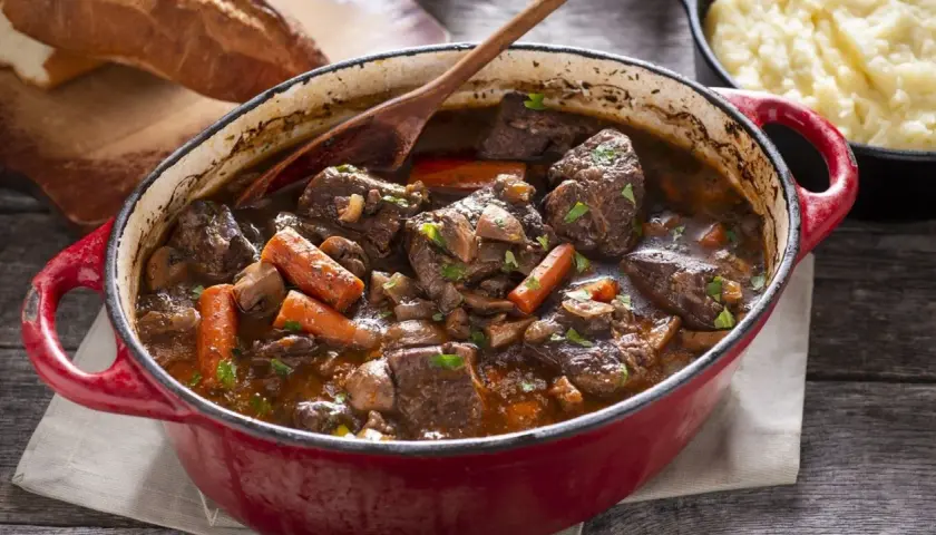 Un invitante spezzatino di manzo in un tegame rosso, con carote, funghi e pane rustico sullo sfondo.