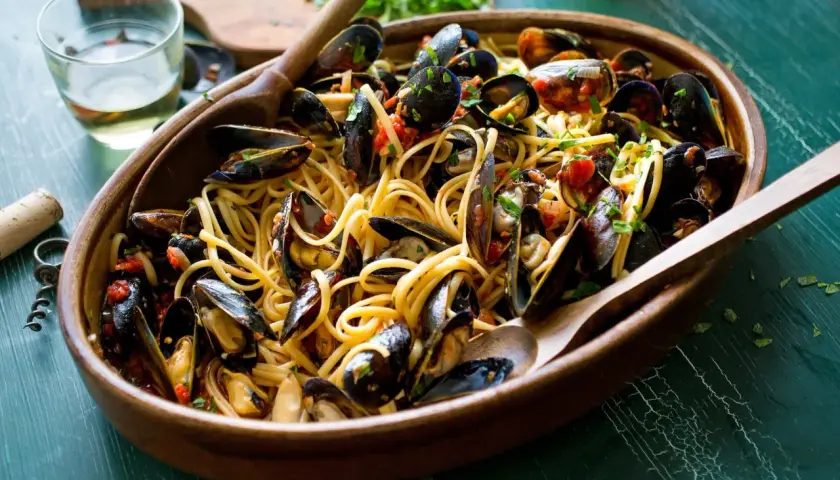 Un invitante piatto di pasta con le cozze, condita con pomodoro e prezzemolo, servita in una ciotola di legno con cucchiai.