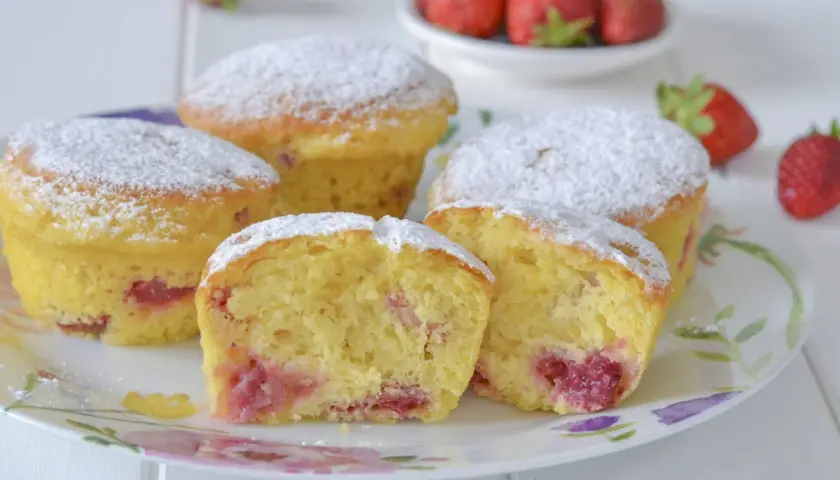 Morbidi muffin alle fragole, cosparsi di zucchero a velo, invitano a un dolce assaggio. Pezzi di fragola succosa si intravedono nell'impasto soffice.