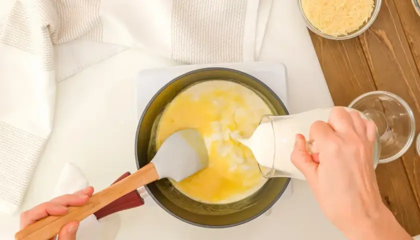 Mani versano latte in una padella con uova e formaggio grattugiato, preparando una deliziosa crema di parmigiano.