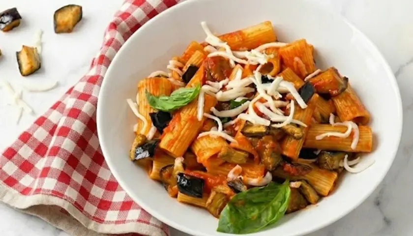 Un piatto di pasta con melanzane, pomodoro e formaggio grattugiato, guarnito con basilico fresco. Accanto, un tovagliolo a quadri rossi e bianchi.