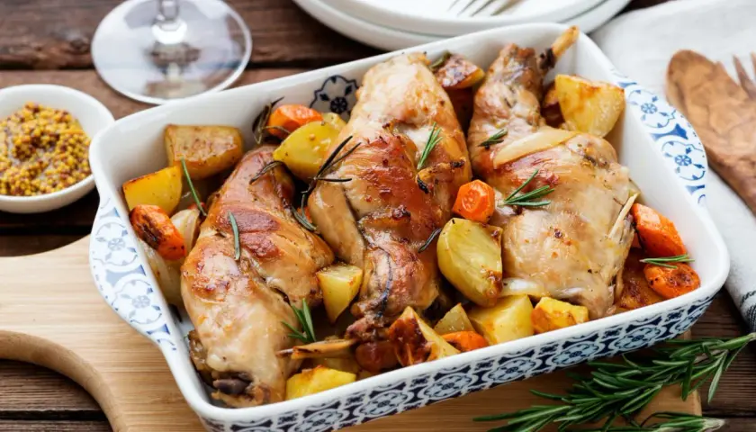 Coniglio al forno dorato con patate e carote, profumato al rosmarino, servito in una pirofila decorata.