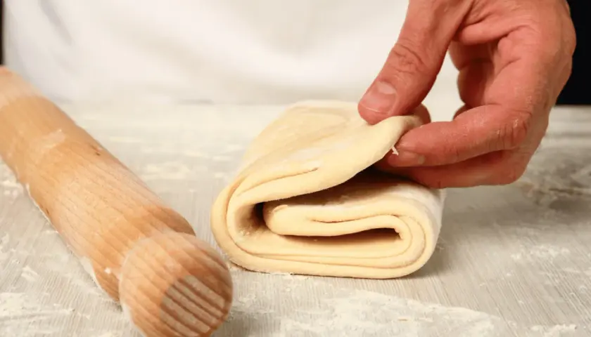 A hand folds layers of puff pastry dough next to a wooden rolling pin on a floured surface.