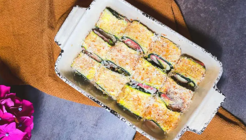 Involtini di zucchine pronti per essere cotti, ripieni e impanati, in una teglia bianca su un tovagliolo arancione.