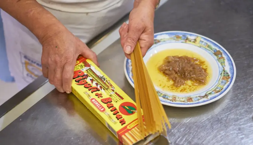 Mani che preparano i bigoli in salsa, con la pasta che esce dalla scatola e la salsa pronta nel piatto.