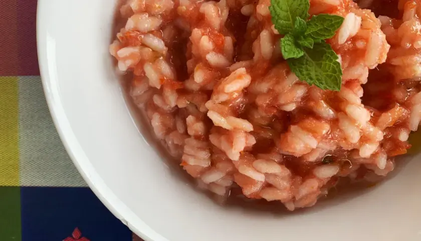 Un cremoso riso al pomodoro, guarnito con una fogliolina di menta fresca, servito in una ciotola bianca su una tovaglia colorata.