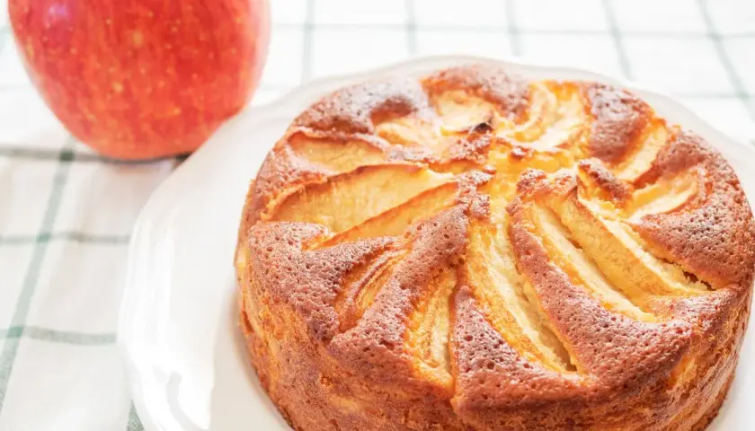Torta di mele con yogurt, dorata e soffice, con mele a vista. Accanto, una mela rossa fresca.