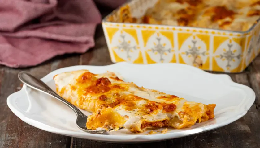 Piatto di cannelloni al forno fumanti, ripieni di carne e ricoperti di besciamella e sugo. Una forchettata mostra il ripieno invitante.