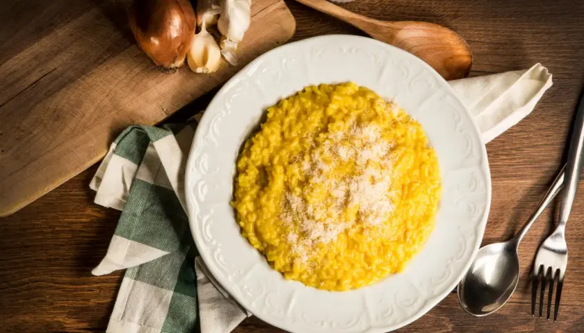 Un cremoso risotto alla milanese, spolverato di parmigiano, pronto per essere gustato. Accanto, cipolla e aglio, ingredienti essenziali.