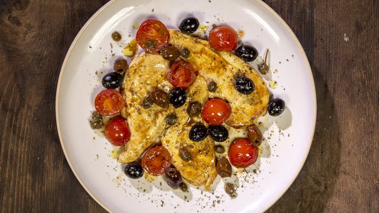Petto di pollo in padella con pomodorini, olive nere e capperi, condito con erbe aromatiche.