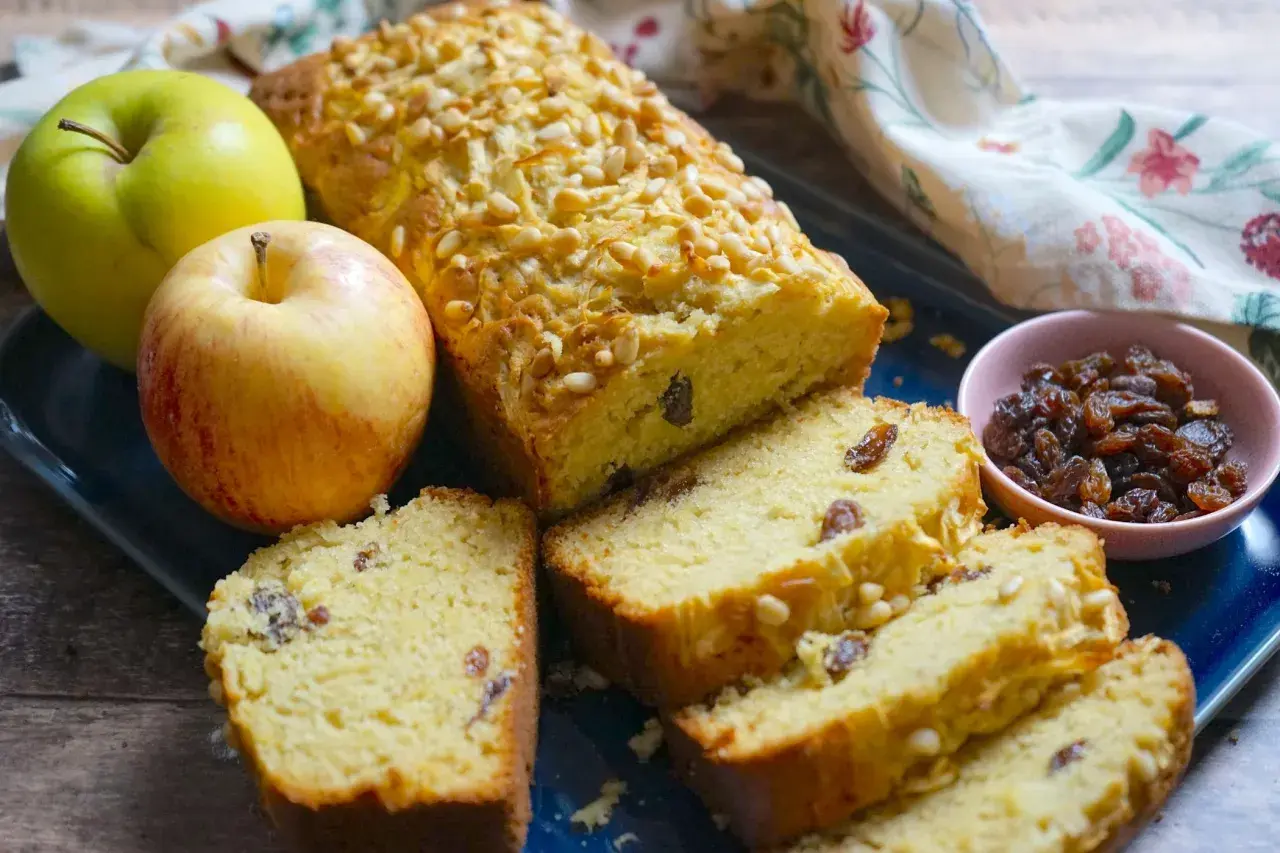 Plumcake alle mele soffice e profumato, con uvetta e pinoli, accompagnato da mele fresche e una ciotolina di uvetta.