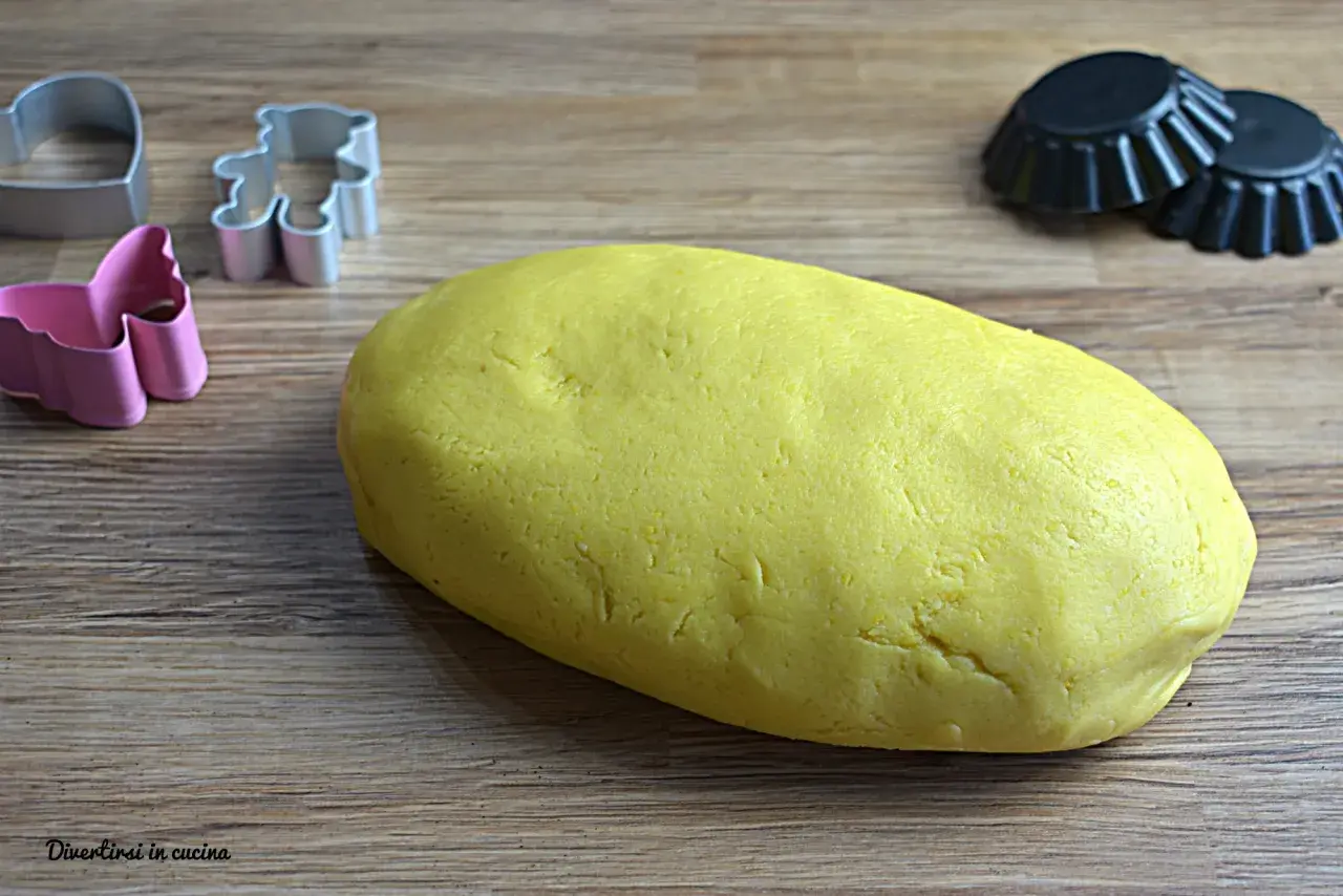 Yellow pasta frolla dough with cookie cutters and tart molds on a wooden surface.