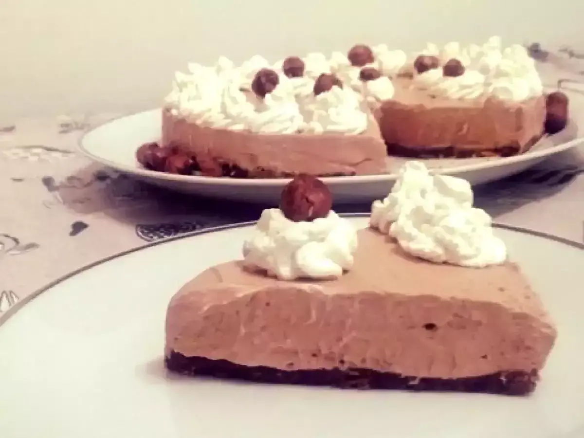 Una fetta di golosa cheesecake alla Nutella, guarnita con panna montata e nocciole intere, su un piatto bianco.
