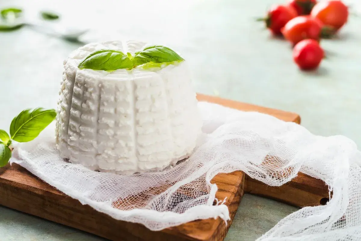Ricotta fatta in casa, fresca e profumata, guarnita con basilico su un tagliere di legno. Pomodorini rossi sfocati sullo sfondo.