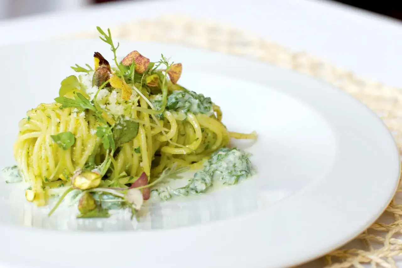 Spaghetti conditi con un cremoso pesto di pistacchi, guarniti con pistacchi interi e erbe fresche.