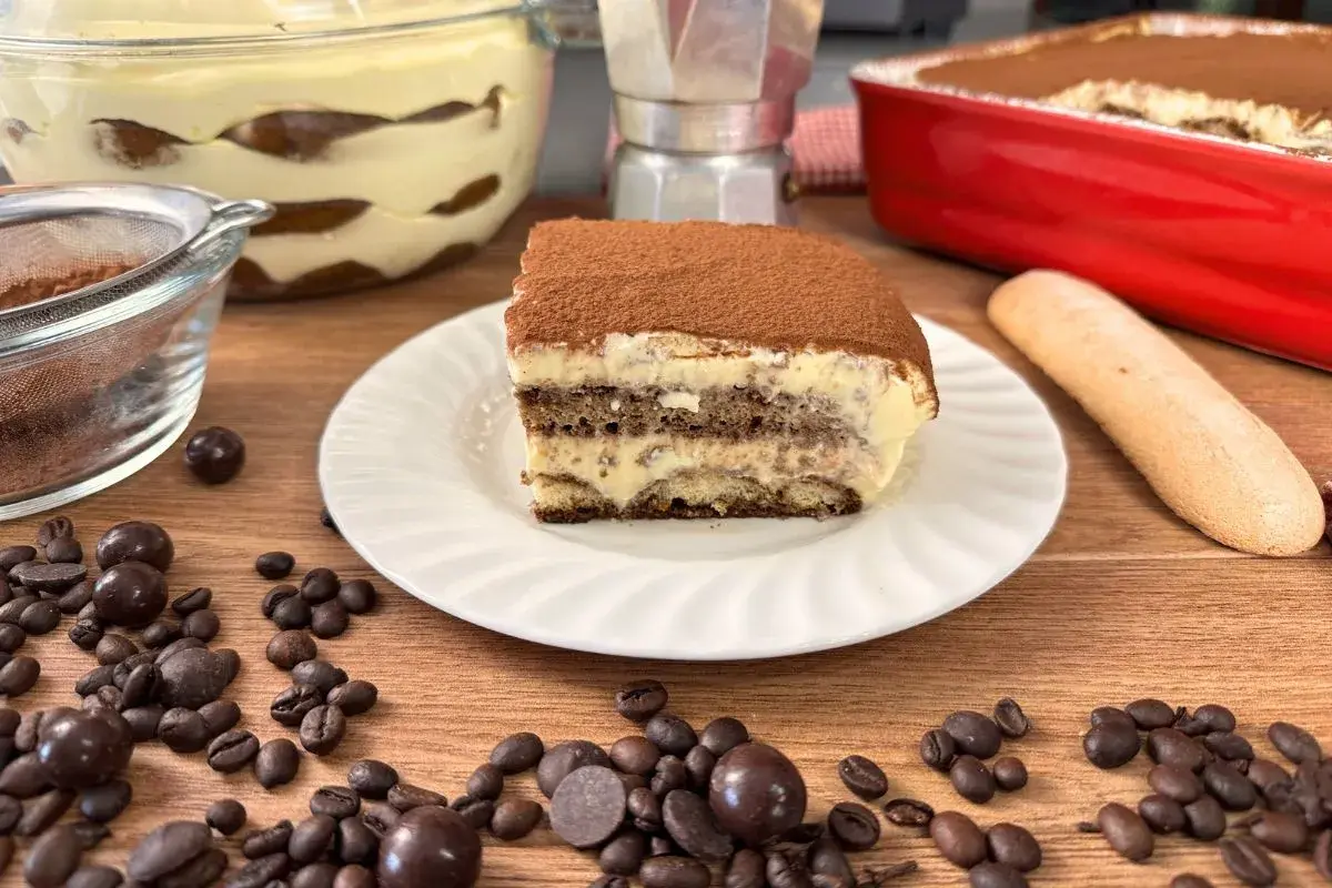 Una fetta di tiramisù con uova pastorizzate, cosparso di cacao, su un piatto bianco, con chicchi di caffè e cioccolatini sparsi.