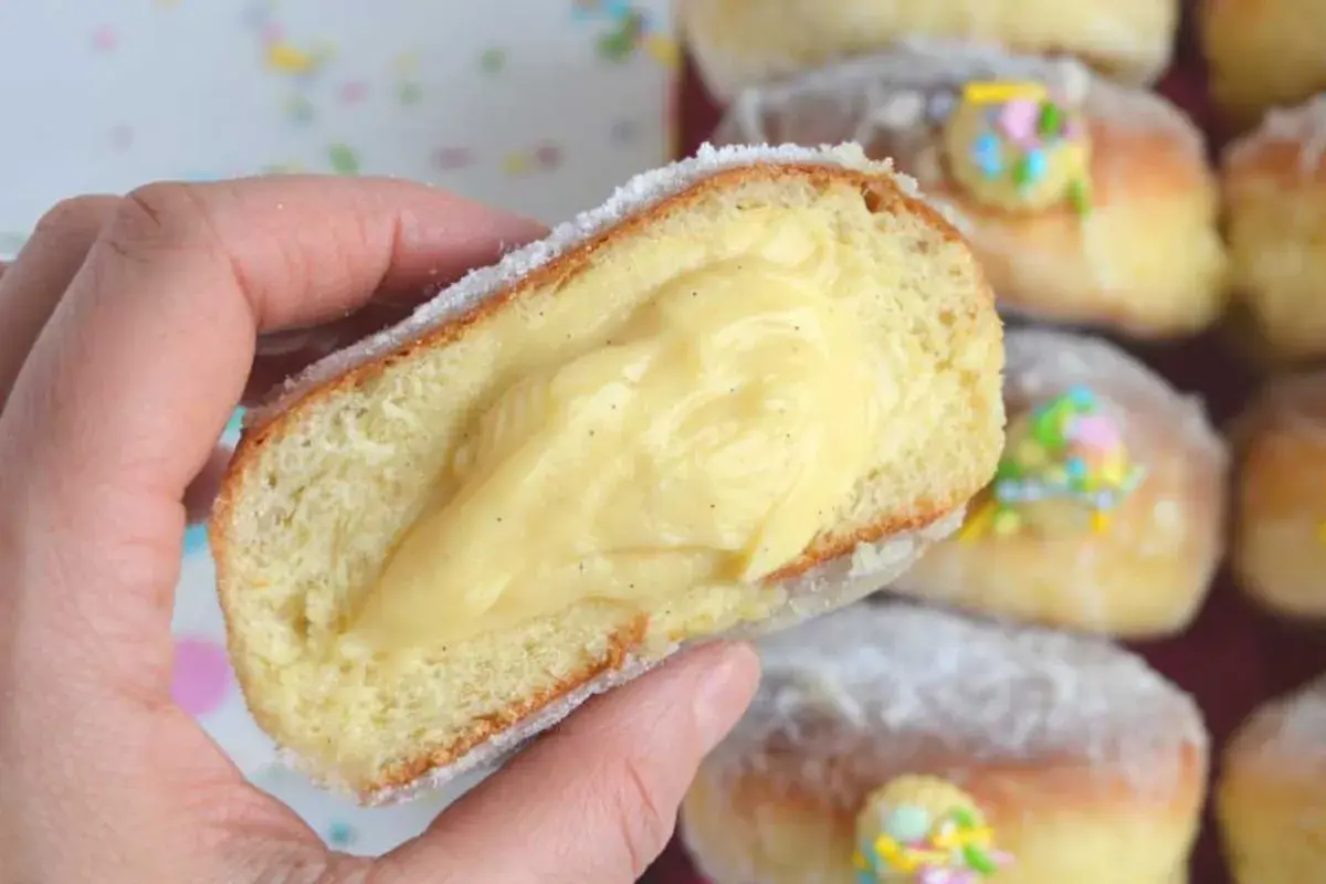 Un bombolone alla crema, soffice e ripieno, viene tenuto in mano. Altri bomboloni alla crema decorati con confettini colorati sono sullo sfondo.