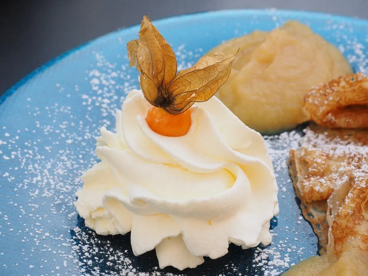 Panna montata soffice con physalis, su un piatto blu cosparso di zucchero a velo, accanto a un dolce che ricorda il pan di spagna.