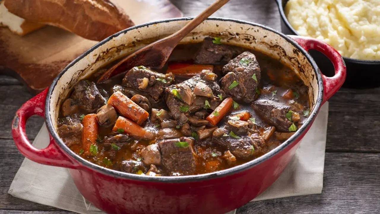 Un invitante spezzatino di manzo in un tegame rosso, con carote, funghi e pane rustico sullo sfondo.