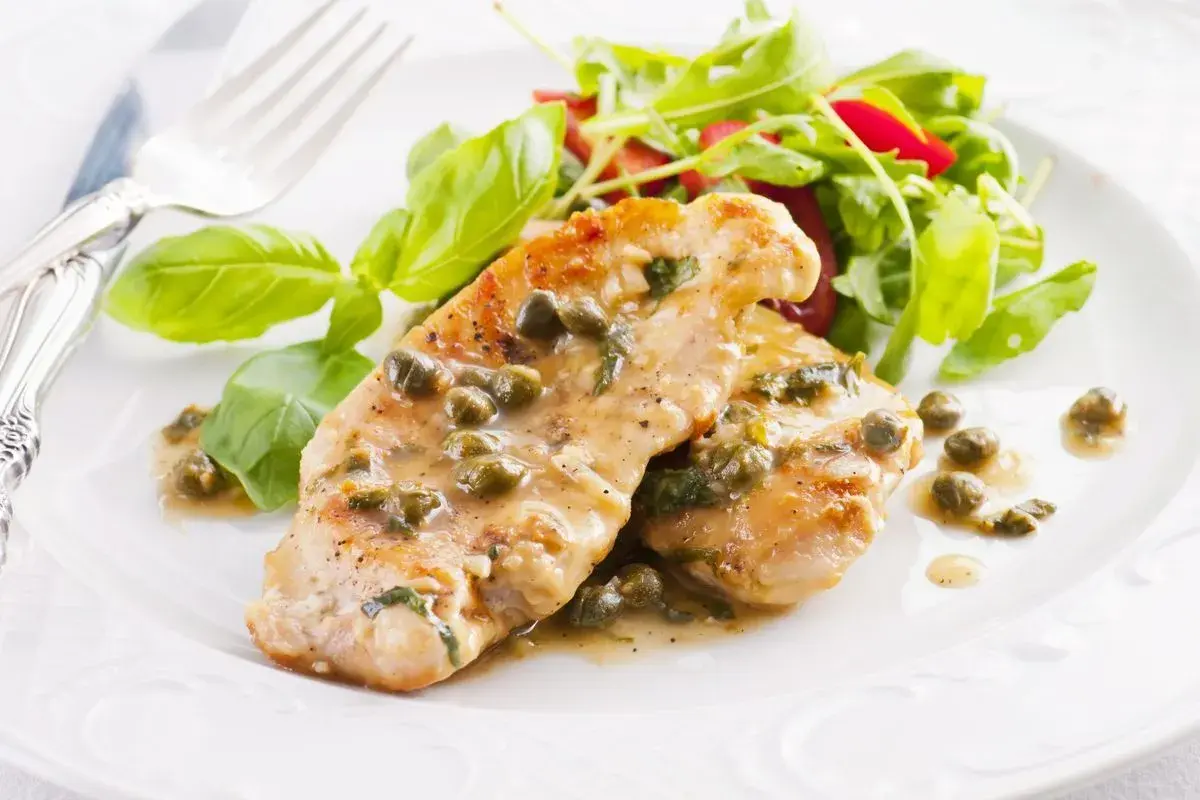Deliziose scaloppine di pollo al limone e capperi, servite con un'insalata fresca e foglie di basilico.