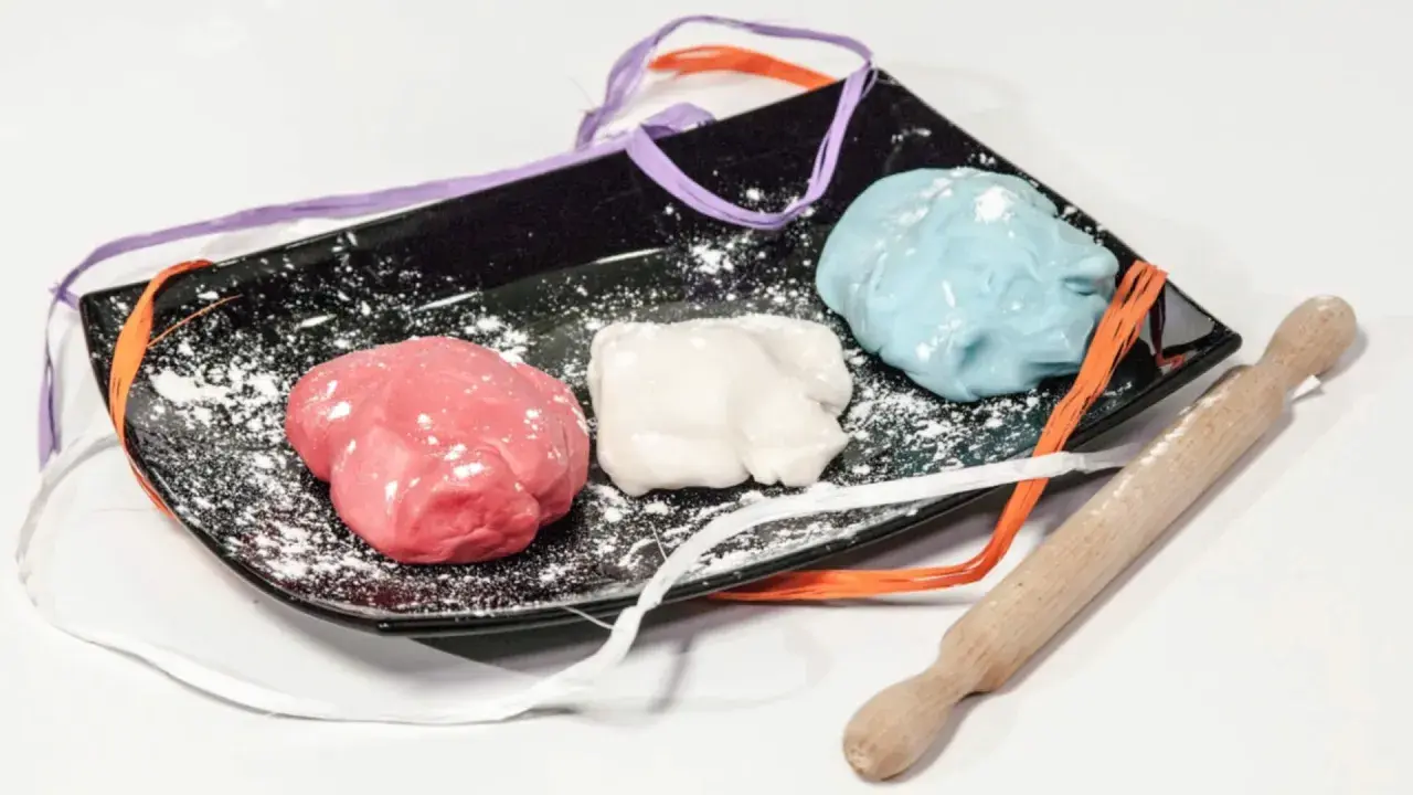 Tre panetti di pasta di zucchero colorata (rosa, bianca, azzurra) su un piatto nero con nastri colorati e un mattarello.