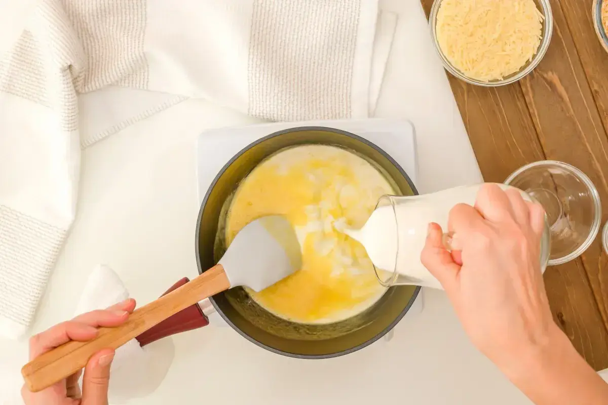 Mani versano latte in una padella con uova e formaggio grattugiato, preparando una deliziosa crema di parmigiano.