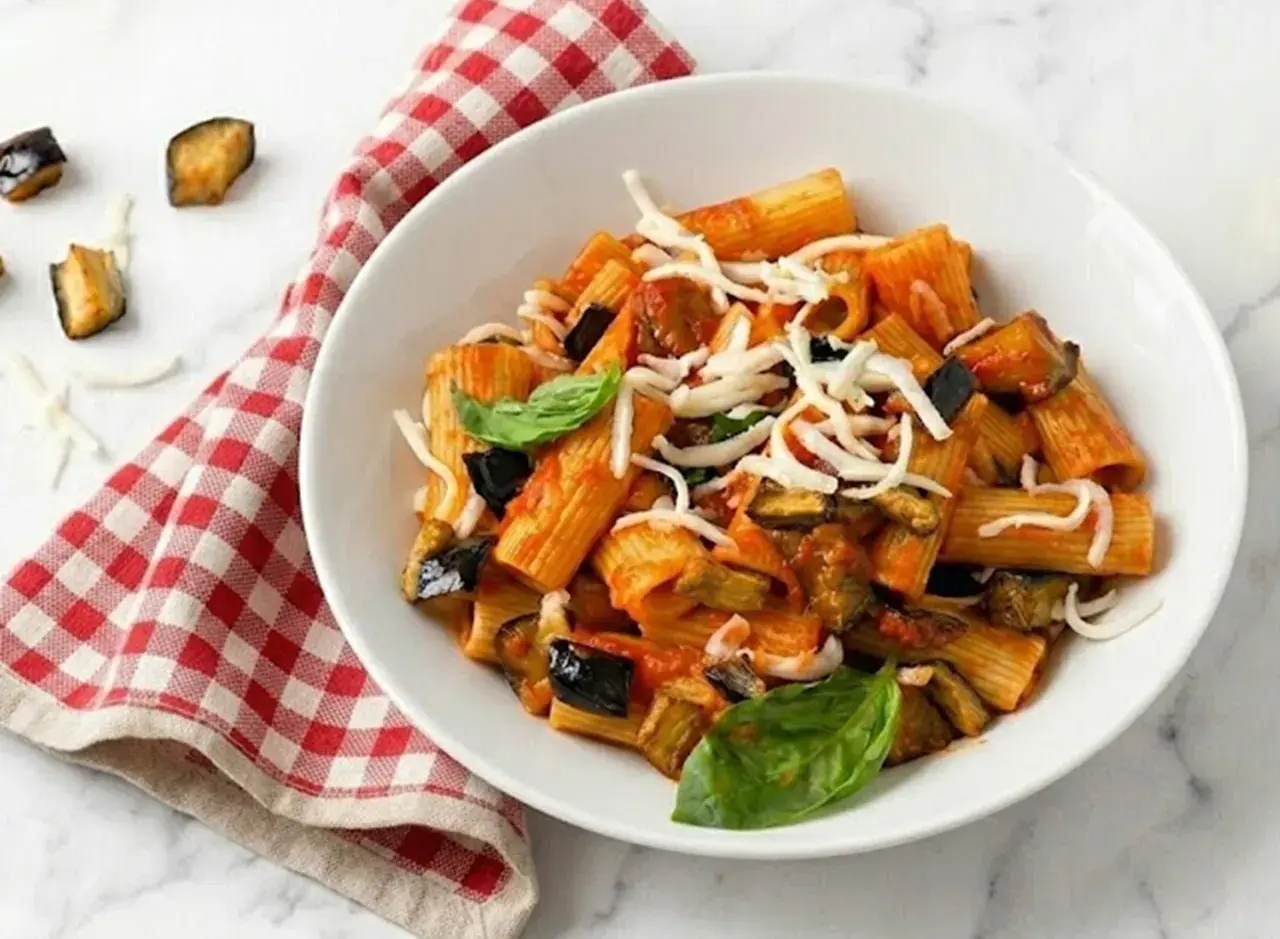 Un piatto di pasta con melanzane, pomodoro e formaggio grattugiato, guarnito con basilico fresco. Accanto, un tovagliolo a quadri rossi e bianchi.