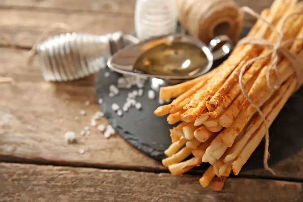 Grissini fatti in casa dorati, legati con spago, su un tagliere con sale grosso, olio e un rotolo di filo metallico.