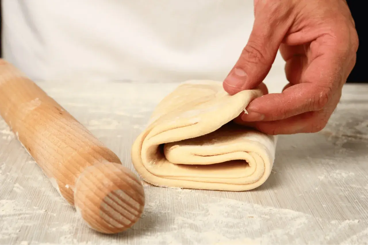 A hand folds layers of puff pastry dough next to a wooden rolling pin on a floured surface.
