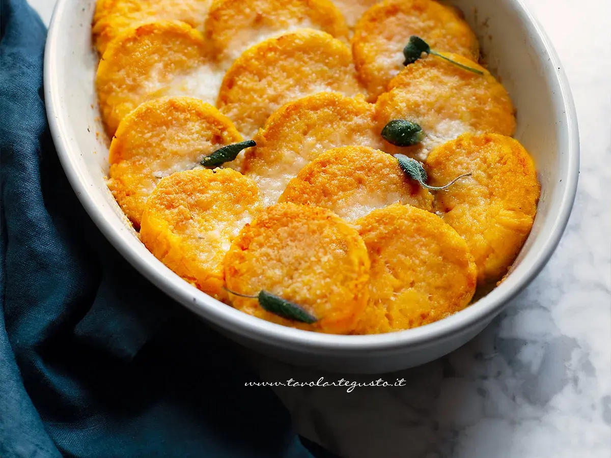 Gnocchi di zucca dorati e gratinati, guarniti con foglie di salvia, in una pirofila bianca.