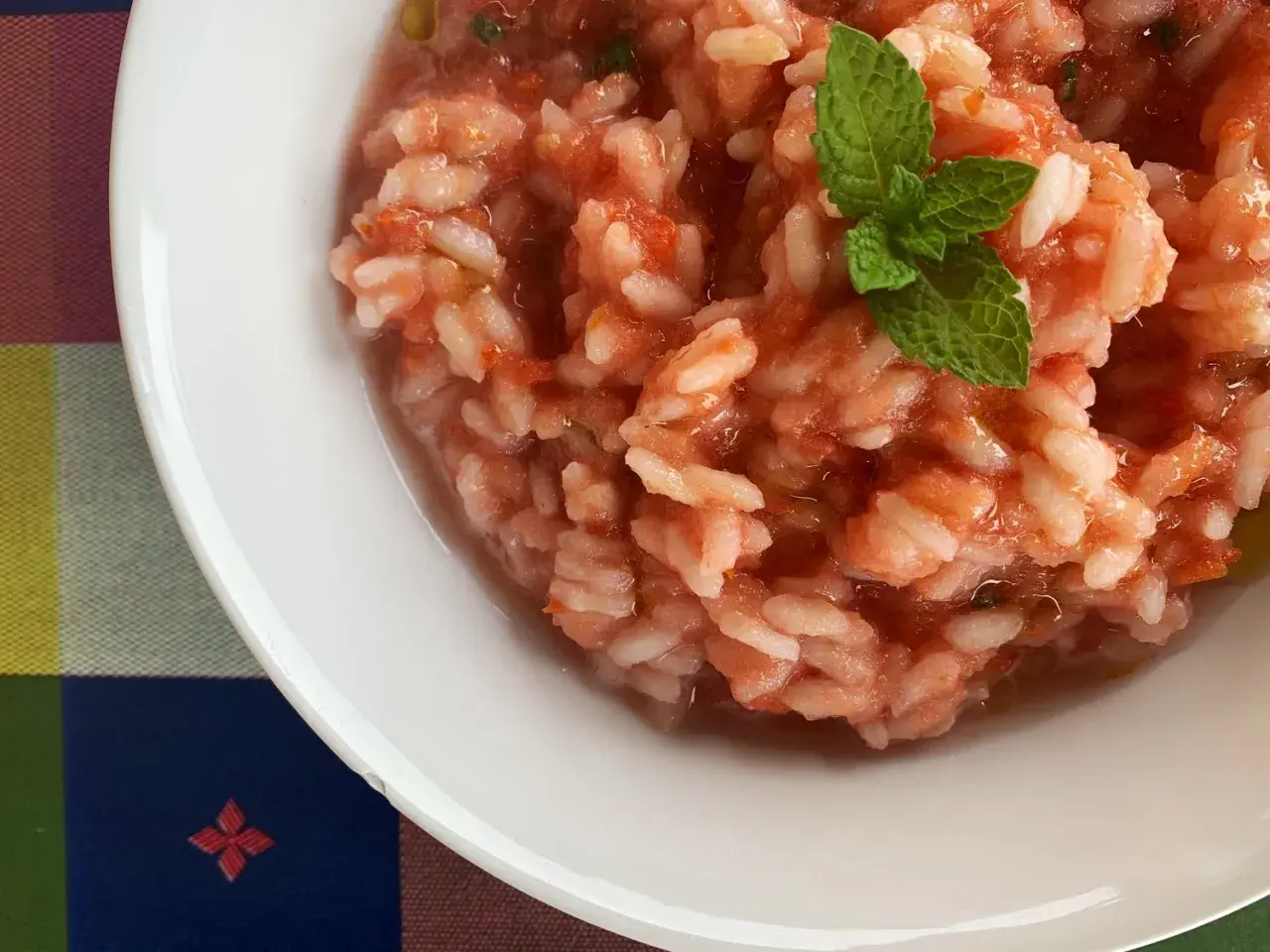 Un cremoso riso al pomodoro, guarnito con una fogliolina di menta fresca, servito in una ciotola bianca su una tovaglia colorata.