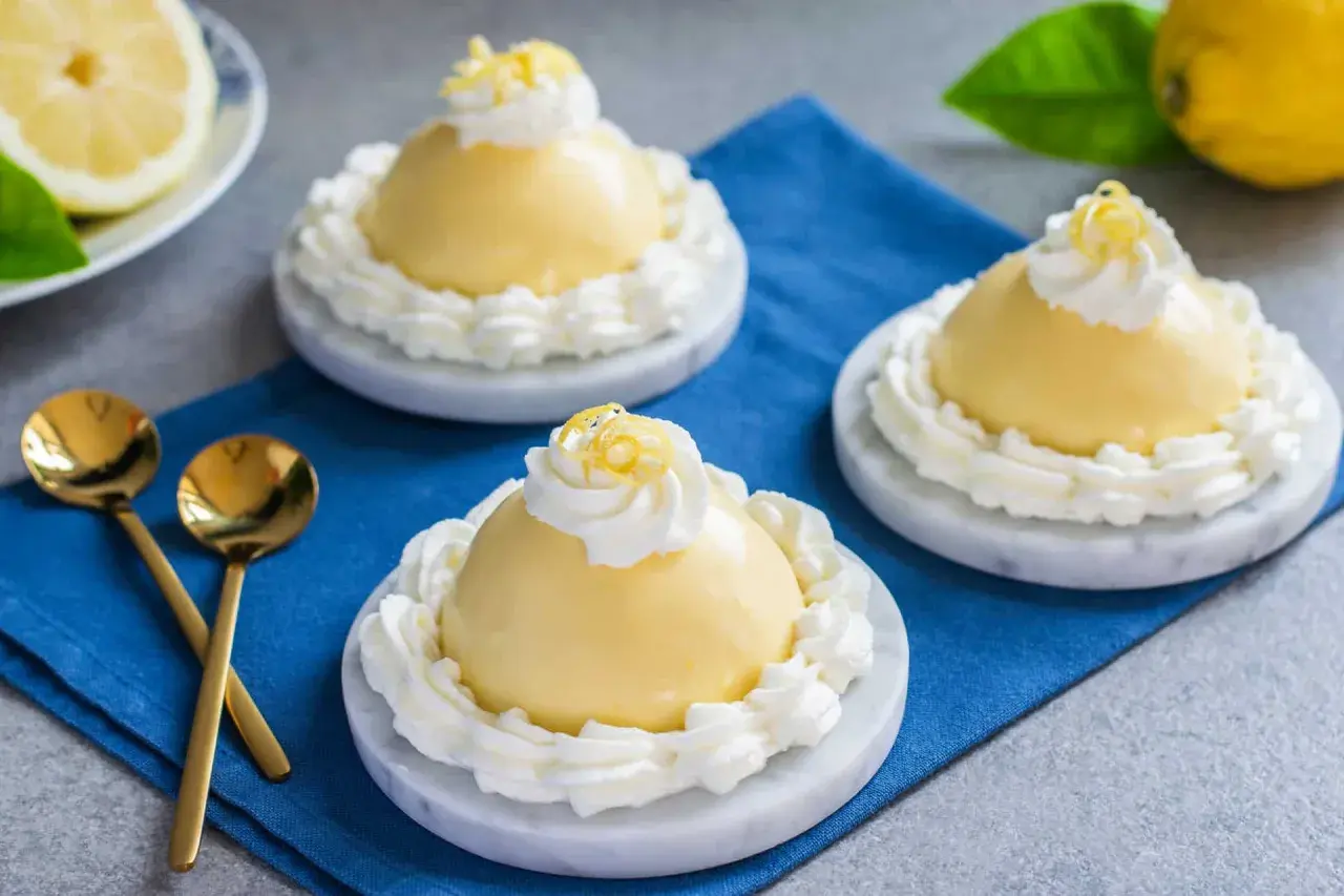 Tre deliziose delizie al limone, guarnite con panna montata e scorza di limone, su piattini di marmo.
