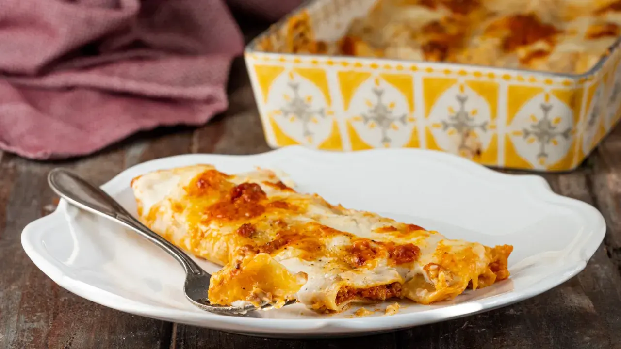 Piatto di cannelloni al forno fumanti, ripieni di carne e ricoperti di besciamella e sugo. Una forchettata mostra il ripieno invitante.
