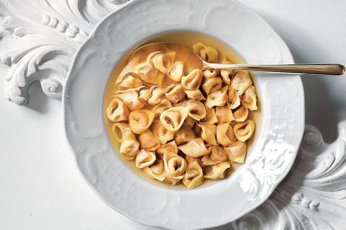 Un piatto di tortellini in brodo, un classico confortante, servito in una ciotola bianca decorata con un cucchiaio dorato.