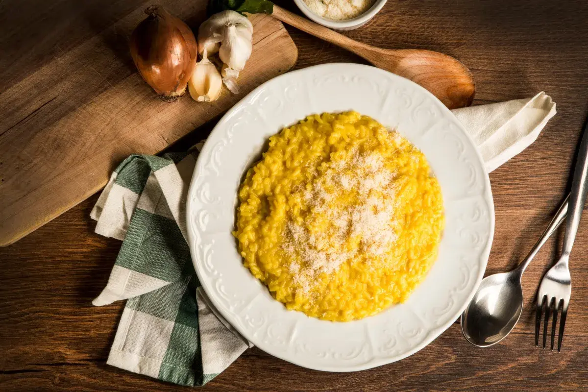 Un cremoso risotto alla milanese, spolverato di parmigiano, pronto per essere gustato. Accanto, cipolla e aglio, ingredienti essenziali.