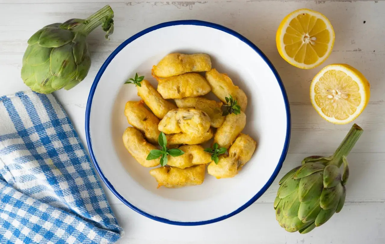 Carciofi fritti dorati, preparati con una pastella croccante, serviti in un piatto bianco con limone e carciofi freschi.
