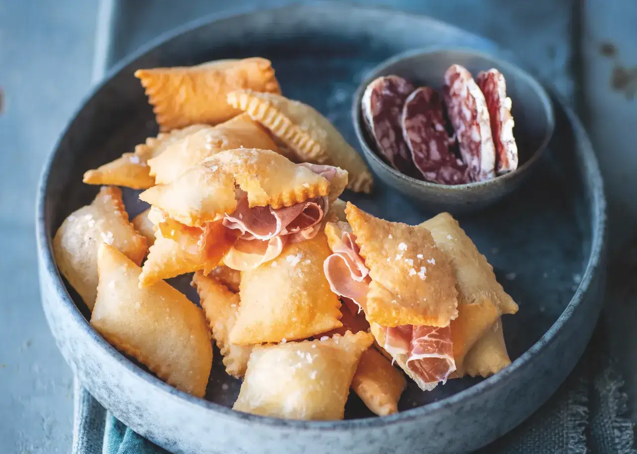Gnocco fritto ricetta: sfogliatine dorate ripiene di prosciutto crudo, cosparse di sale grosso, servite in un piatto con salumi affettati.