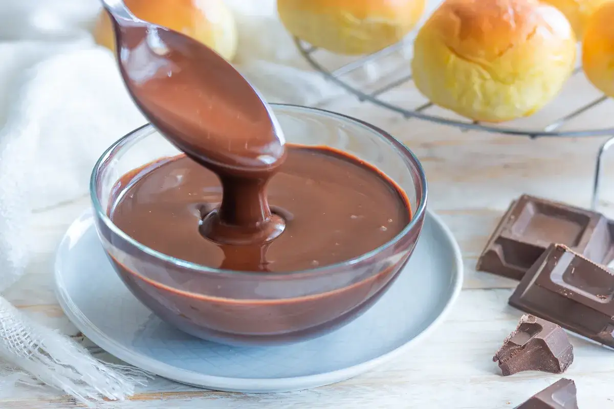 Cucchiaio versa lucida ganache al cioccolato in una ciotola di vetro. Sullo sfondo, panini dorati e pezzi di cioccolato.